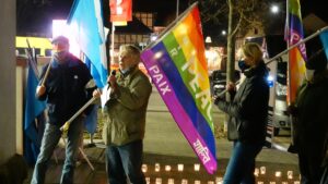 Wolfgang Lieberknecht, chapter coordinator for the WBW Wanfried chapter in Germany, speaks into a microphone at an outdoor rally for peace, holding a rainbow-colored flag with peace written on it in multiple languages. Other members of the rally stand to his left and right, each holding peace flags and wearing COVID masks.