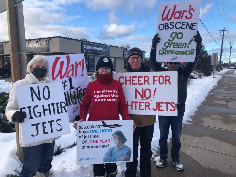 Activists stand outside MP Terry Dowdall's office holding signs protesting Canada's planned fighter jets purchase