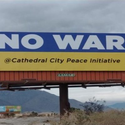 A billboard with the colors of the Ukraine flag. The top blue stripe has huge white lettering saying "NO WAR." The bottom yellow stripe has the words "@Cathedral City Peace Initiative." Behind the billboard is a stormy sky with mountains in the distance.