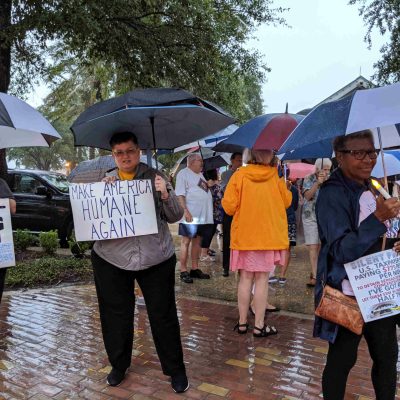 A Florida chapter member stands in the rain holding an umbrella and a sign reading "Make America Humane Again", in a crowd of other protesters holding signs and umbrellas.