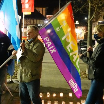 Wolfgang Lieberknecht, chapter coordinator for the WBW Wanfried chapter in Germany, speaks into a microphone at an outdoor rally for peace, holding a rainbow-colored flag with peace written on it in multiple languages. Other members of the rally stand to his left and right, each holding peace flags and wearing COVID masks.